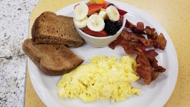 A plate of fluffy scrambled eggs, crisp bacon and wheat toast beside a bowl of fresh fruit