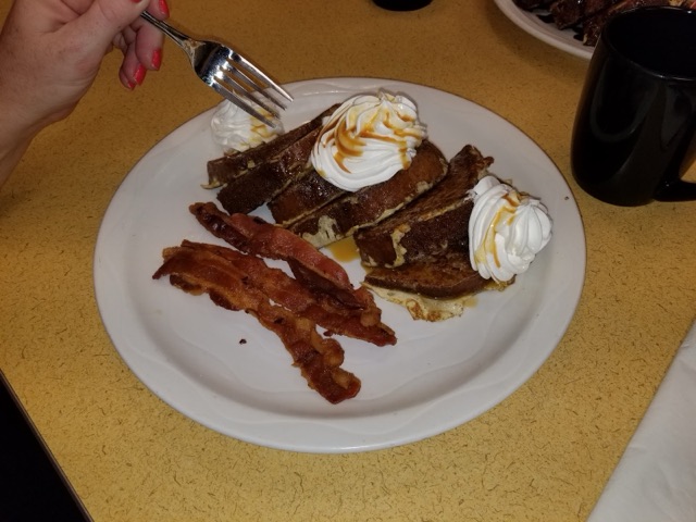 Thick slices of French toast topped with whipped cream and a caramel drizzle, with crisp bacon alongside