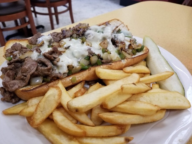 A Philly cheesesteak of steak, peppers, onions and melted cheese on a toasted roll, with steak-cut fries and a pickle